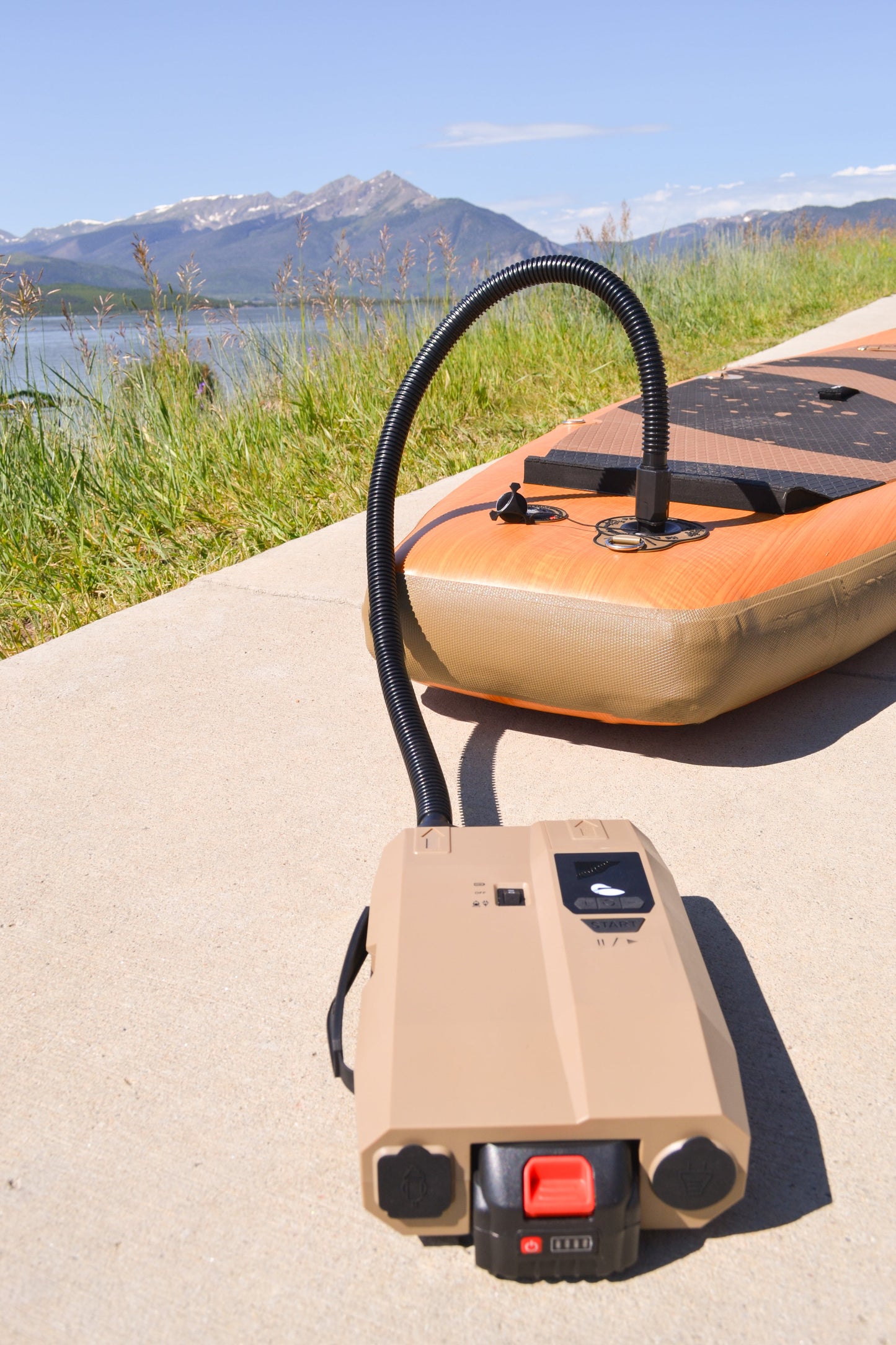 Tan inflator with black hose plugged into a paddle board on a sidewalk by the lake.