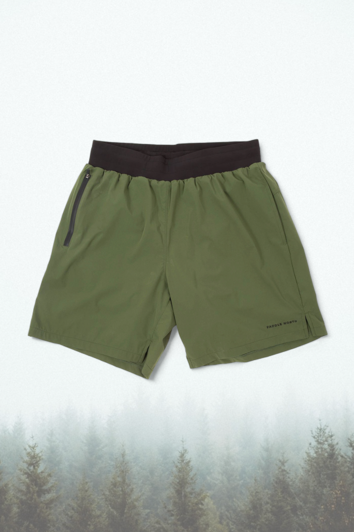 Green shorts with a black elastic waistband against an evergreen tree background.