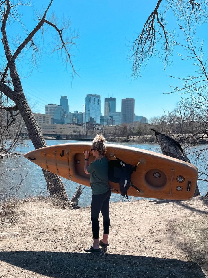 Person holding an inflatable kayak by a river.