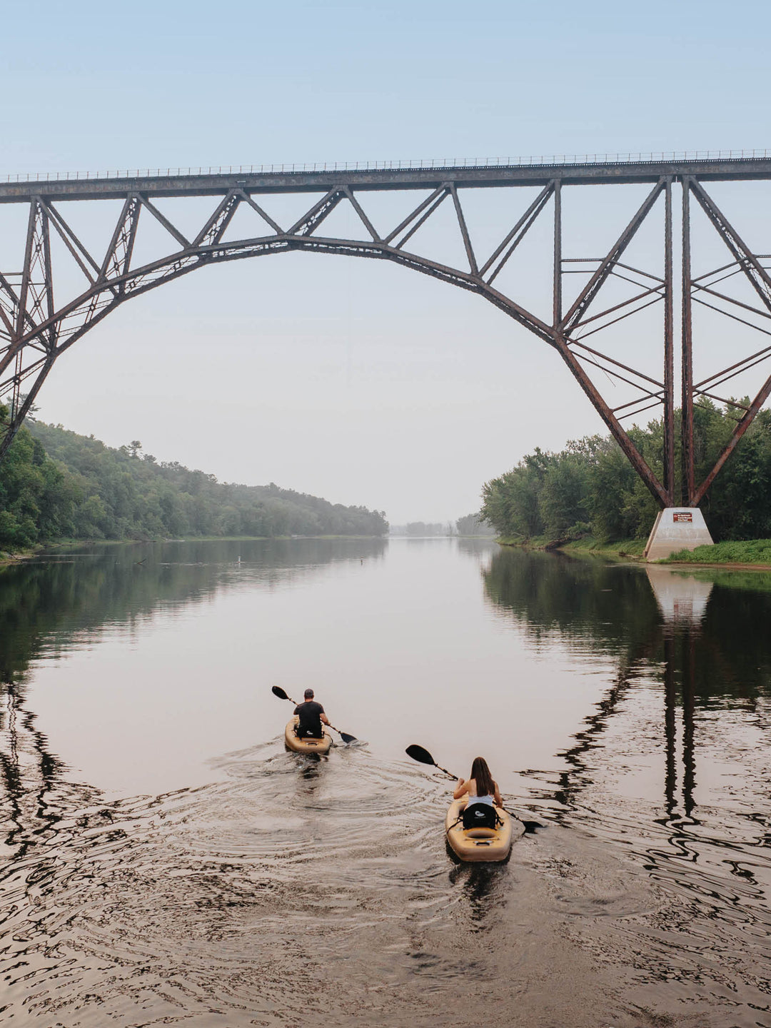 Two people kayaking on a river heading towards a bridge.