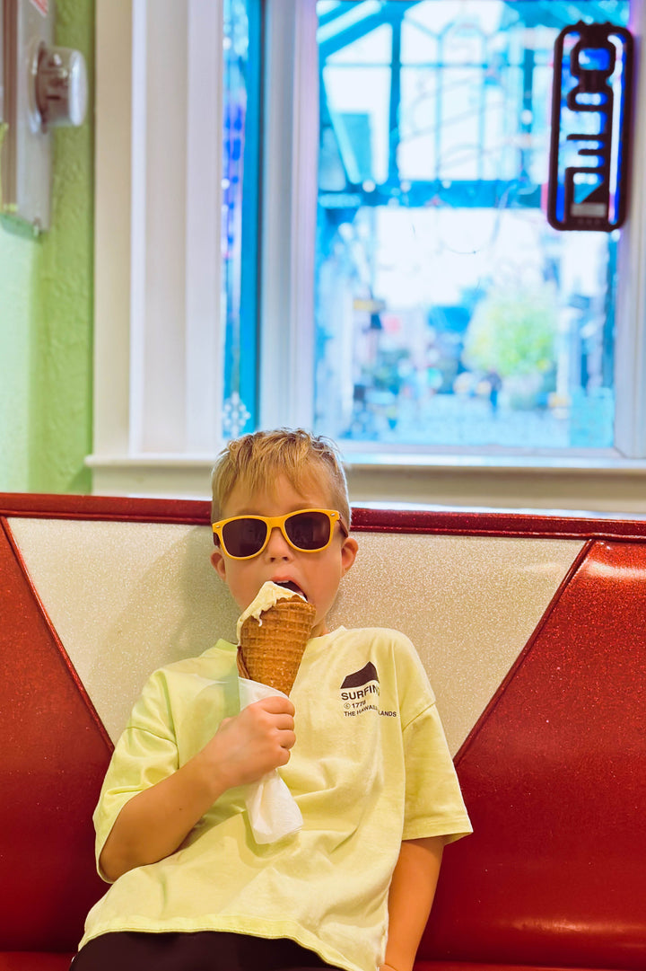 Little kid eating ice cream wearing yellow framed sunglasses with wood temple piece.